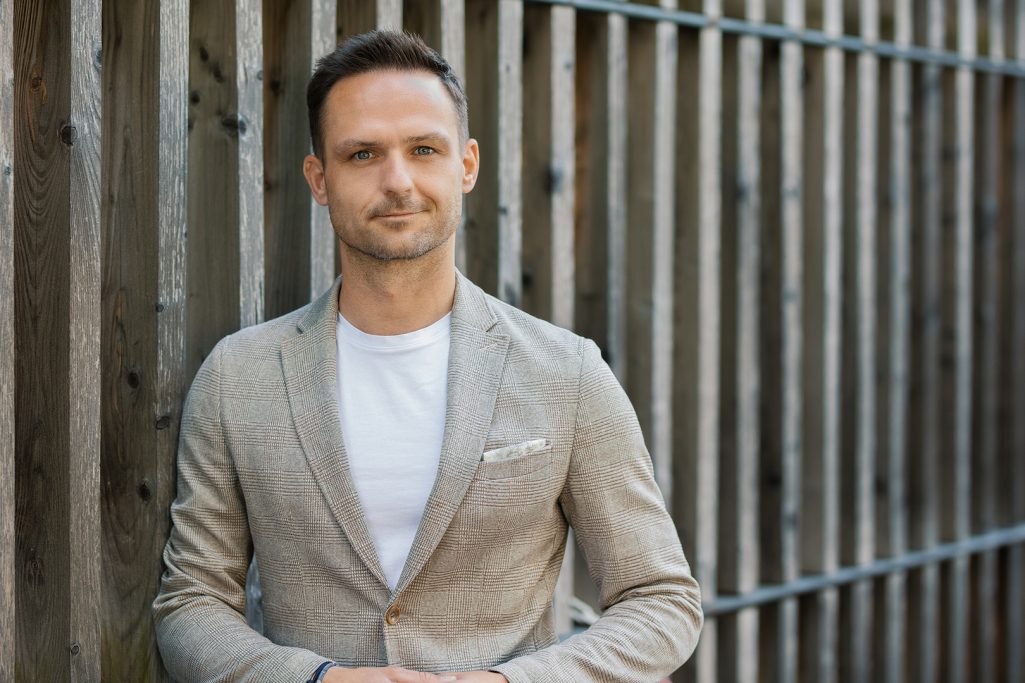 Mann in heller Jacke steht vor einem Holzzaun, lächelt leicht in die Kamera.