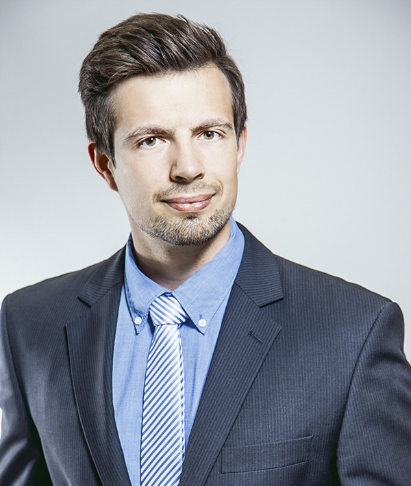 Ein gut gekleideter Mann in Anzug mit Krawatte und lächelndem Gesichtsausdruck.