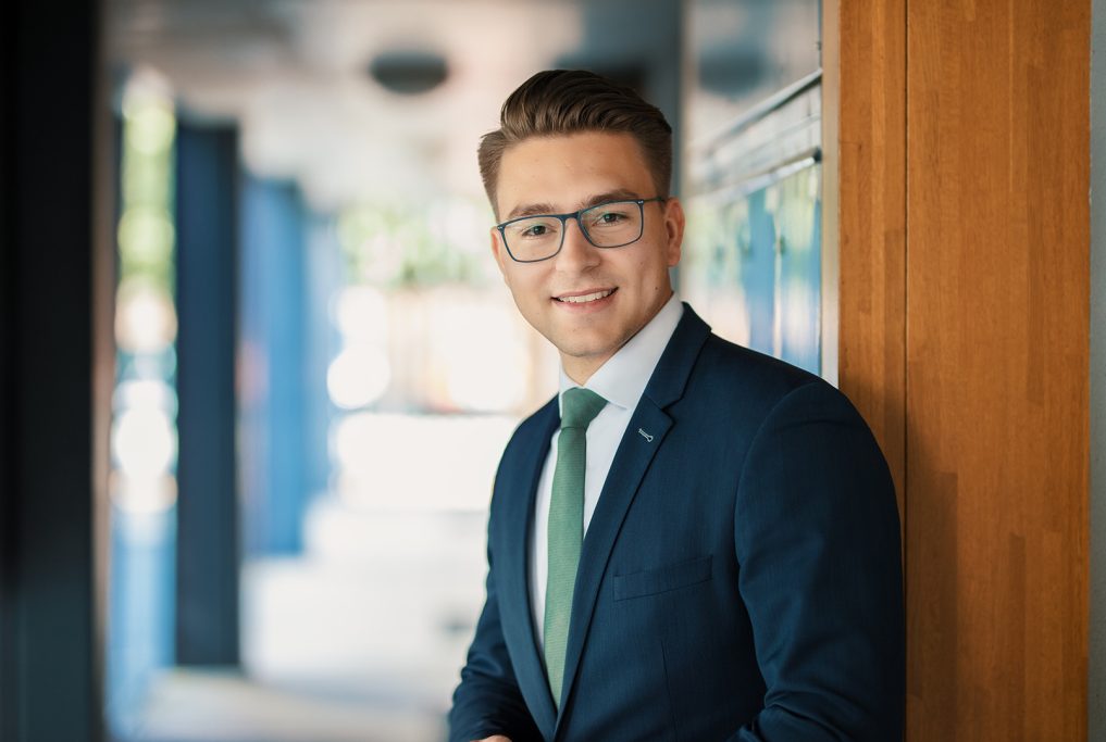 Ein junger Mann in Anzug und Krawatte lächelt selbstbewusst vor einem modernen Hintergrund.