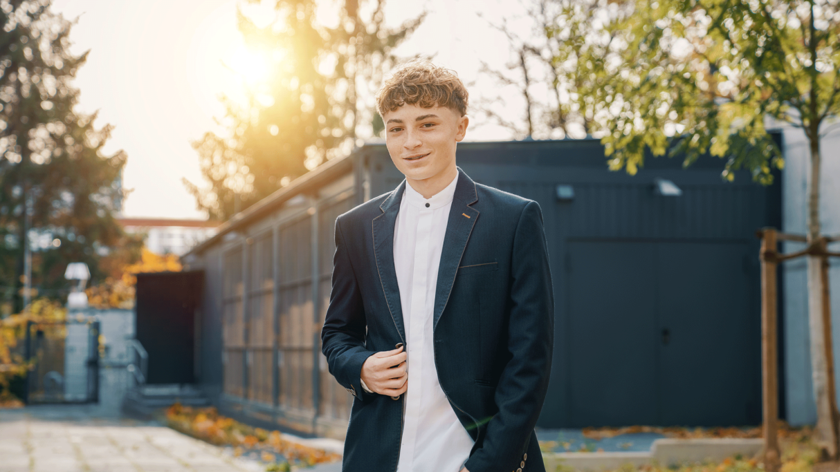 Junger Mann in Anzug lächelt, umgeben von Bäumen und Sonnenlicht.