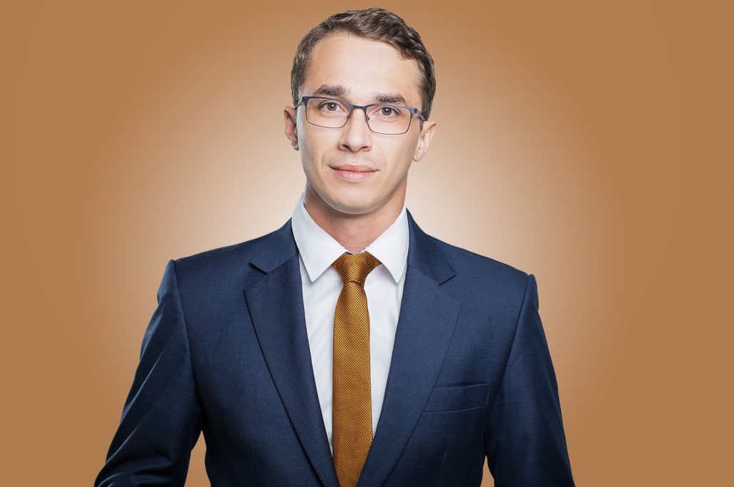 Junger Mann in Anzug mit Krawatte, lächelt in neutralem Hintergrund.