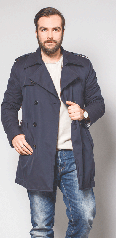 Mann in blauem Trenchcoat und Jeans, posiert vor neutralem Hintergrund.